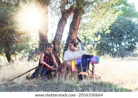 Two hikers, man and woman resting under the shade of a tree and chatting animatedly after a long walk. 