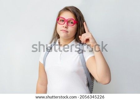 Portrait of little schoolgirl in pink glasses with backpack holding index finger up