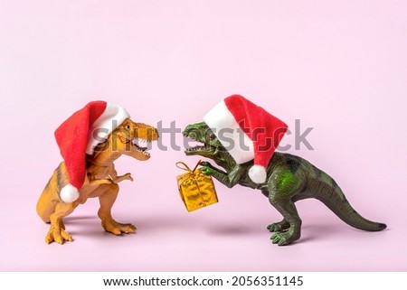 Two dinosaurs Rex in red Santa Claus hat holds golden gift box in its paws on pink background New Years Eve or Christmas Eve Art holiday card Creative idea for Merry xmas concept
