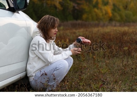 A pretty young woman in a white hoodie sits near a white car with a thermos in her hands and drinks hot tea on a warm autumn day against a background of yellow foliage. Selective focus. Portrait