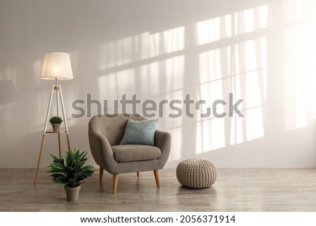 Comfortable retro armchair with pillow, luminous lamp and plant in pot, ottoman on floor on gray wall background in living room. Cozy interior in Scandinavian style, contemporary design with furniture