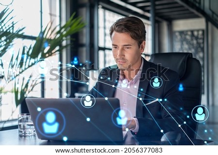 Handsome businessman in suit at workplace working with laptop to hire new employees for international business consulting. HR, social media hologram icons over office background