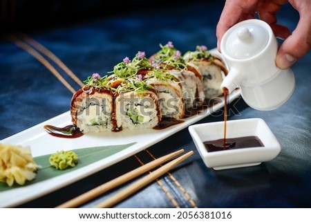 sushi roll philadelphia with eel on a white long plate on a blue background hand pours sauce into a gravy boat
