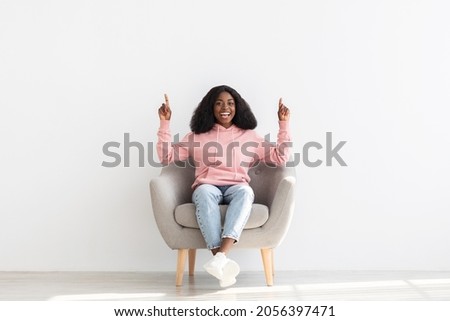 Positive african american young lady sitting in arm chair over white wall at home, pointing up at copy space for text or advertisement, happy black woman recommending something exciting Royalty-Free Stock Photo #2056397471