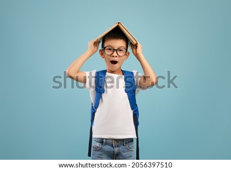 Back To School. Funny Excited Asian Schoolboy Keeping Book On Head And Looking At Camera, Nerdy Korean Male Child Wearing Eyeglasses And Backpack Standing Isolated Over Blue Background, Copy Space