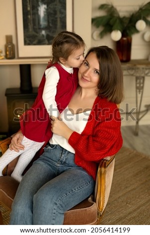 A girl kisses her mother sitting in an armchair by the fireplace on Christmas Eve. Children's love for their parents. Casual clothes in red colors. 