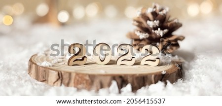 New year 2022. Numbers 2022 on wooden stand on beige pastel blurred background with decorative fir trees, cone, snow and lights. Christmas greeting card. Banner