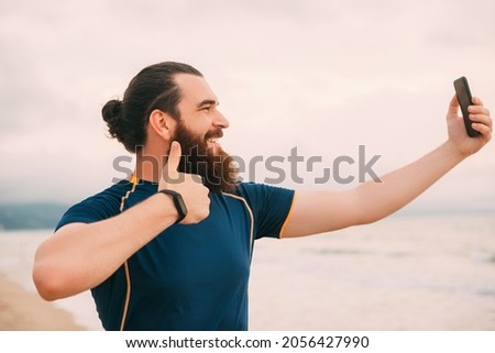 Bearded hipster man is taking a selfie with thumb up after running on the beach.