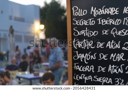 Restaurant menu list written in spanish (translation from the top: chicken, iberian pork, cheese, tuna tartare, ham, tuna, tongue). Granada, Spain. Blurred people in the background.