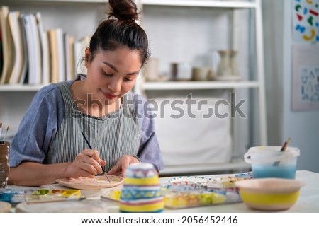 Asian woman learning color painting her self-made pottery at home. Confidence female enjoy hobbies and indoors leisure activity handicraft ceramic sculpture and painting workshop at pottery studio. Royalty-Free Stock Photo #2056452446