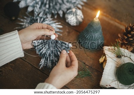 Hands holding stylish handmade angel from blue wrapping paper on background of rustic wooden table with paper stars, candle. Atmospheric moody image, nordic style. Winter holidays preparation
