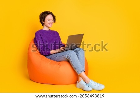Full length photo portrait of woman working on laptop sitting in orange beanbag chair isolated on vivid yellow colored background