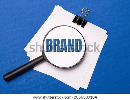 On a blue background, white sheets under a black paper clip and on them a magnifying glass with the text BRAND
