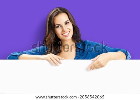 Happy smiling brunette young woman in blue clothing, standing behind, peeping from blank banner or showing advertising mock up signboard with copy space for some text, over violet purple background.