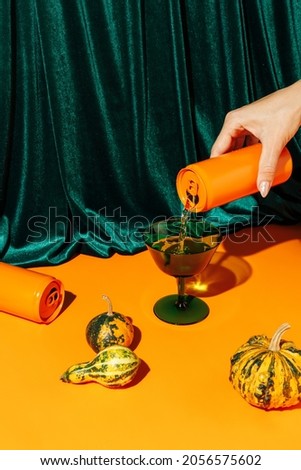 Orange pumpkins and a woman's hand pouring a beverage drink from an orange can against plush velvet curtain background. Creative Halloween and Thanksgiving concept. Contemporary fall still life idea.