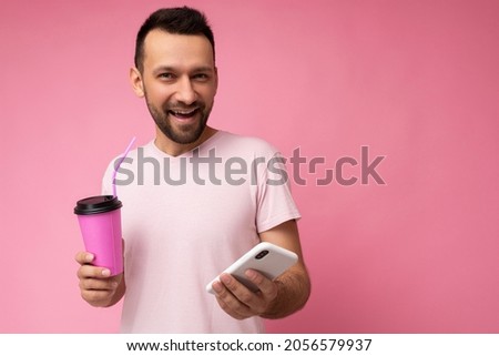 Photo of joyful smiling handsome young unshaven brunette man with beard wearing everyday light pink t-shirt isolated over pink background holding and using mobile phone surfing on the internet online