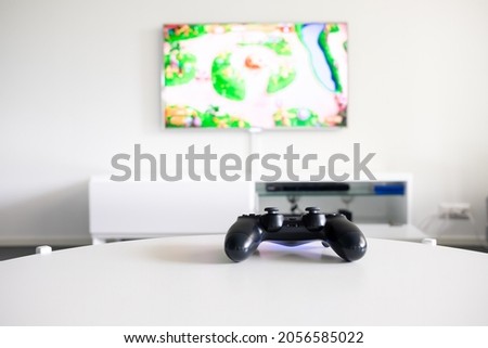 A view of a logo free, black videogame controller on a white table. Tv is in the background.