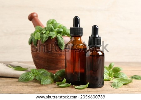 Bottles of basil essential oil near mortar with leaves and pestle on wooden table