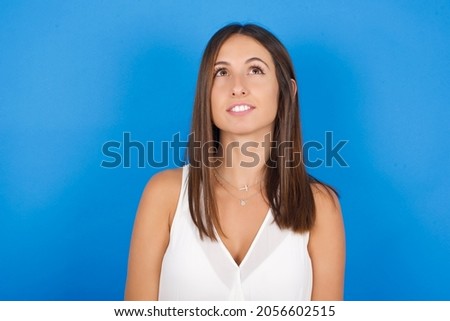 Young european brunette woman wearing white T-shirt on blue background looking up as he sees something strange.