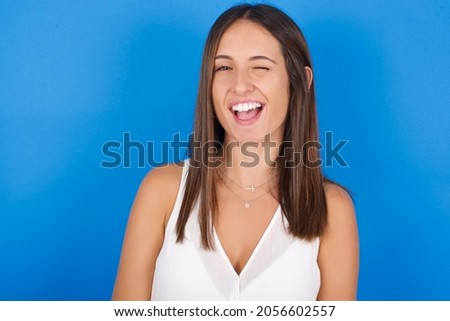Young european brunette woman wearing white T-shirt on blue background blinking eyes with pleasure having happy expression. Facial expressions and people emotions concept.