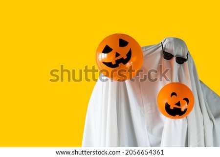 Person in Halloween costume of ghost with sunglasses and Jack-o'-lantern balloons smiling Royalty-Free Stock Photo #2056654361
