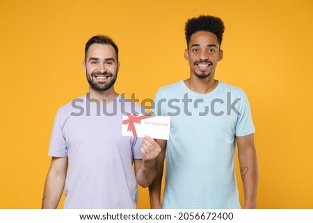 Smiling young two friends european african american men 20s wearing violet blue casual t-shirts hold gift certificate looking camera isolated on bright yellow colour wall background studio portrait