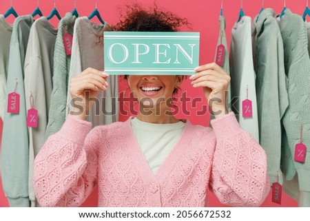 Young happy female costumer woman wear sweater stand near clothes rack with tag sale in store showroom cover face with card sign with open title text isolated on plain pink background studio portrait