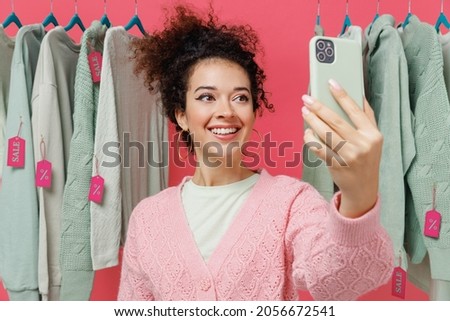 Young female costumer woman in sweater stand near clothes rack with tag sale in store showroom do selfie shot on mobile cell phone post photo on social network isolated on plain pink background studio