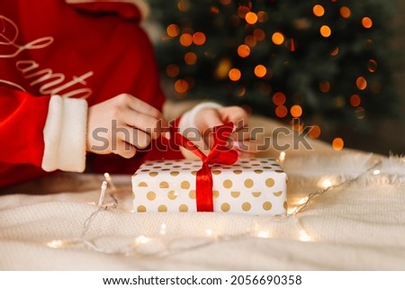 Preparation for the Christmas holiday. Human hands with manicure hold a gift box with a festive decor against a background of light and bokeh on New Year's Eve at home. Selective focus