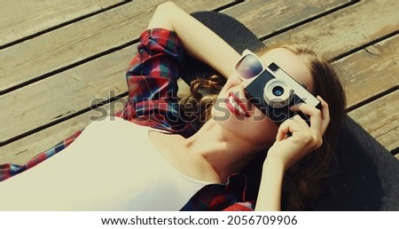 Portrait of happy smiling young woman photographer with vintage film camera lying on a skateboard outdoors