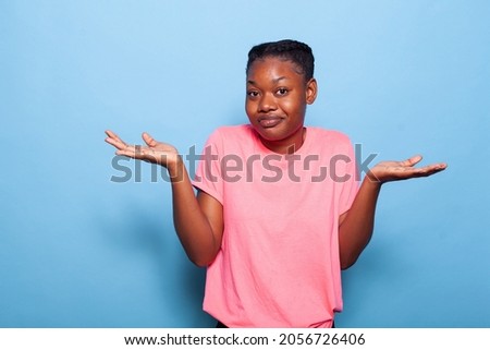 Portrait of confused african american teenager posing in pink t-shirt on a blue background in studio making uncertain facial expression. Young woman shrugging shoulders. I dont know concept Royalty-Free Stock Photo #2056726406