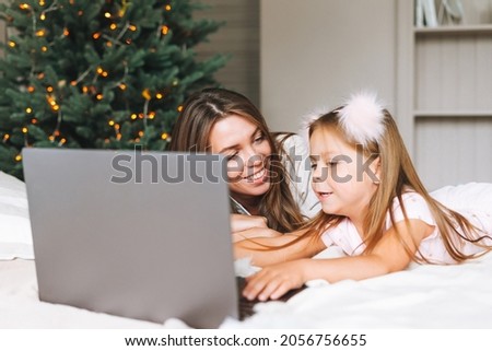 Little girl with mother using laptop on bed in room with Christmas tree at home
