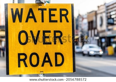 A yellow and black road sign featuring the message “Water Over Road”