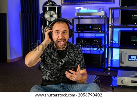 Middle-aged man audiophile listens to music with headphones. Home acoustics, floor-standing speakers stand side by side. Emotions from listening. Amplifier, turntable - rich stereo system. Royalty-Free Stock Photo #2056790402