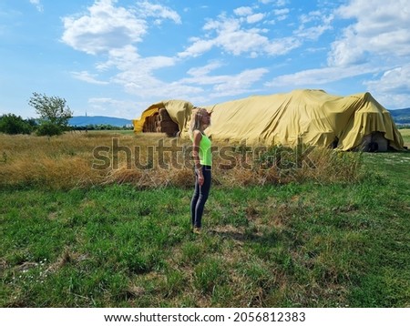 young woman posing near big rectangular straw stacks covered by sail. girl looking aroud. nature summer agriculture wallpaper.  picture of haystacks or straw bales. 
