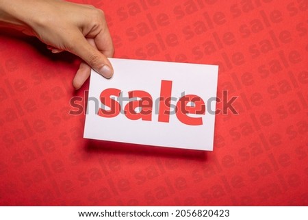 female hand holds a piece of paper with the inscription sale over strong red background cardboard with sale written on to promote special sales