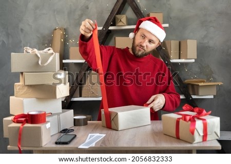 a man in a Santa hat prepares Christmas gifts in a warehouse for delivery and ties a red ribbon. Cardboard boxes on the table and the businessman is packing. New Year sale concept Royalty-Free Stock Photo #2056832333