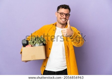 Young caucasian man making a move while picking up a box full of things isolated on purple background with thumbs up because something good has happened
