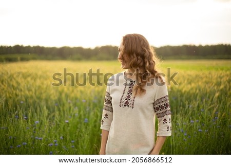 The girl is standing in the rye field. The young girl is in the green rye field.  Woman is wearing Ukrainian national clothes, embroidered shirt. Smiling young lady with blue eyes, wavy blond hair.