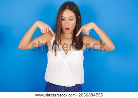 Amazed Young european brunette woman wearing white T-shirt on blue background  points down with fore fingers, opens mouth being shocked. Advertisement concept.