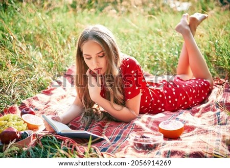 Picnic scene: pretty girl lying on a plaid and reading book in grove. Outdoor portrait of beautiful young woman in red dress