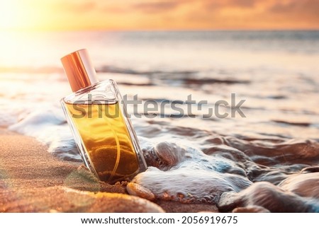 A glass perfume bottle with a golden lid on a sandy beach in surf. Close-up, view from the lower angle. Sunset in the background. Copy space. Perfume advertising concept. Royalty-Free Stock Photo #2056919675