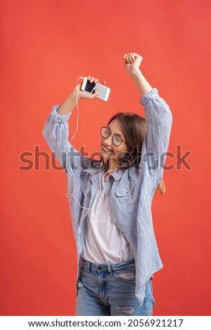 Charming girl in casual clothes and earphones dancing, moving hands, smiling, enjoying the moment isolated on red background.