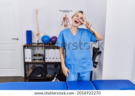 Young physiotherapist woman working at pain recovery clinic doing peace symbol with fingers over face, smiling cheerful showing victory 