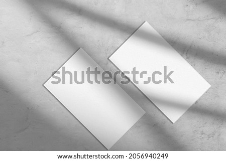 Two empty white rectangle a4 poster, business card or flyer mockups lying diagonally with abstract window shadow overlay on trendy gray concrete background.