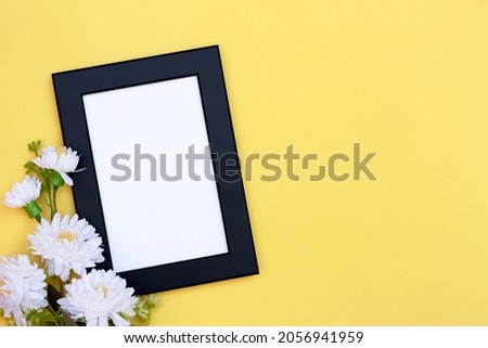 Blank photo frame decorated with white flowers on yellow background. Mockup, top view. Empty space for text.