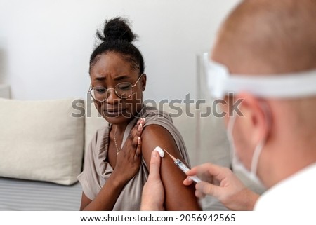 Young African American female patient sitting in a medical clinic and is being given the Covid 19 vaccine in her shoulder by a male Caucasian doctor, wearing protective face mask.