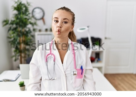 Young caucasian woman wearing doctor uniform and stethoscope at the clinic making fish face with lips, crazy and comical gesture. funny expression. 