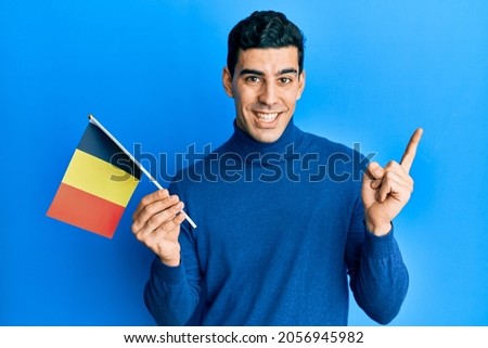 Handsome hispanic man holding belgium flag smiling happy pointing with hand and finger to the side 