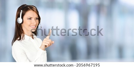 Call center service. Customer support female phone sales operator in white confident cloth, headset showing pointing clicking at copy space, imaginary or text, standing over blurred office background.